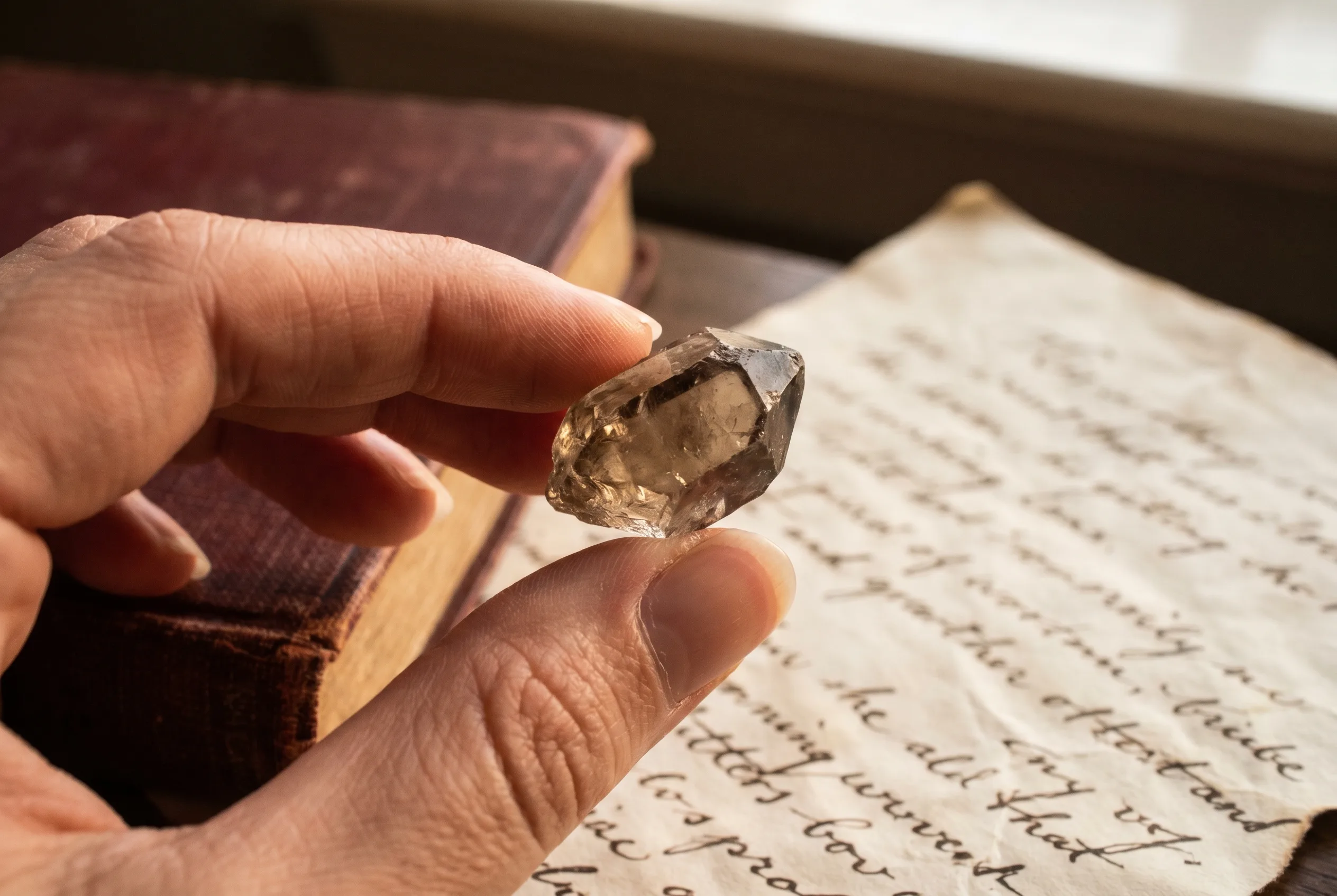 Macro photograph of fingertips holding a single rough smoky-quartz crystal in warm window light, with the corner of a clothbound book and a handwritten parchment out of focus behind.